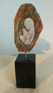 Een verbeelding van een baby in de baarmoeder. De baby is zonder olie waardoor hij licht van kleur is gebleven. De baarmoeder en alles eromheen is wel met olie gedaan waardoor dit bruin/groen is met strepen erdoor. De andere kant van het beeld is een bolle buik (niet te zien). Het is opgezet op een kleine zwarte stenen sokkel en zweeft iets erboven op een pin.