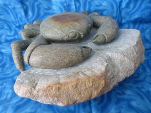 Een krab zit op een rots. Het geheel is gemaakt uit 1 steen. De krab is iets donkerder dan de rots door olie en was. Bovenop de rug van de krab zit een bruine vlek, de krab zelf is verder grijzig met witte strepen en wat bruine vlekjes. De poten van de krab zijn open gewerkt waardoor het lijkt dat hij los op de rots zit. De rots is witgrijs met aan de voorkant een bruine rand.